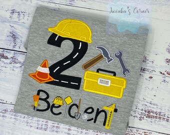 May include: A grey t-shirt with a black number "2" with a white road design, a yellow construction hat, a yellow toolbox, a hammer, a wrench, and an orange traffic cone. The text "Be" and "dent" are written in black with a yellow construction hat, a shovel, and a ruler.