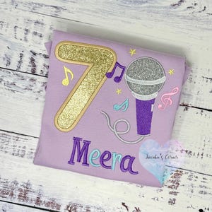 May include: A lilac-colored t-shirt featuring a gold glitter number 7, a silver glitter microphone, and musical notes. The name "Meera" is embroidered in purple and teal. A heart-shaped logo is in the bottom right corner.