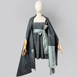 May include: A dark gray sleeveless dress featuring a floral pattern in muted tones, paired with a matching shawl and a wide, light sage green sash tied in a bow at the waist. The dress has a square neckline and a gathered skirt that falls above the knee.