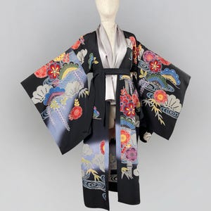 Puede incluir: Un kimono negro con coloridos estampados florales y de olas. El kimono presenta mangas largas y un cinturón negro a juego. El forro interior es de color gris claro. Las mangas y la parte inferior del kimono tienen un efecto degradado.
