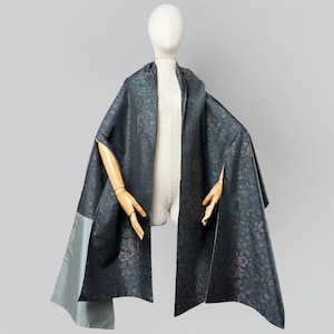 May include: A dark charcoal gray cape featuring an intricate floral pattern in muted tones of blue and purple. The garment has a structured silhouette with wide, open arm slits and a contrasting solid light gray panel on the lower left side.