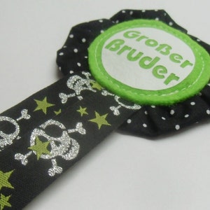 Könnte beinhalten: Schwarz-weiße Stoffrosette mit grünem Besatz und weißem Text "Großer Bruder". Die Rosette ist an einem schwarzen Band mit silbernen Totenköpfen und Knochen und goldenen Sternen befestigt.