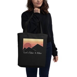 Puede incluir: Bolsa de tela negra con un gráfico de puesta de sol y montaña y el texto "Let's Take A Hike".