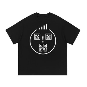 Op de afbeelding: Een zwart T-shirt met korte mouwen en een grote witte afbeelding op de borst. Het ontwerp bestaat uit een cirkelvormige omtrek met twee vierkante QR-code ogen, een uitroepteken als neus en een QR-code mond, met daarboven een signaalbalkicoon.