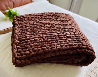 Può includere: Una coperta a maglia grossa piegata, di un ricco colore marrone caldo. La coperta è realizzata con filato spesso, creando un aspetto strutturato e accogliente. La coperta è appoggiata su un letto bianco, suggerendo un ambiente camera da letto confortevole.