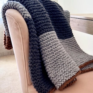 May include: A chunky knit throw blanket featuring horizontal color-blocked stripes in dark charcoal gray, light gray, and a warm brown tone. The blanket has a thick, textured garter stitch pattern and is draped over the back of a light pink chair.