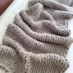 May include: A chunky knit blanket in a neutral gray color. The blanket is made of thick, soft yarn and has a textured appearance. It is draped over a white surface, creating a cozy and inviting look. Perfect for home decor.