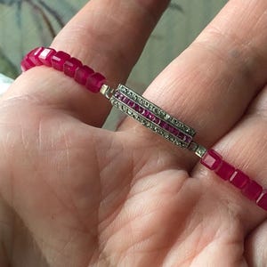 Puede incluir: Primer plano de un collar con un cierre plateado y una hilera de pequeñas cuentas cuadradas de color rubí. El cierre es rectangular y presenta una fila de piedras de color rubí y piedras más pequeñas y transparentes.