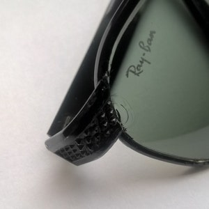 May include: Black Ray-Ban sunglasses with green lenses. The sunglasses have a textured design on the arms.