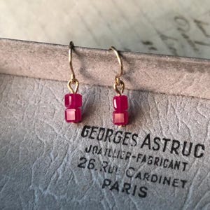 Può includere: Orecchini pendenti con ganci color oro, ciascuno con due perline quadrate fucsia impilate. Gli orecchini sono esposti su una superficie grigia strutturata con il testo "GEORGES ASTRUC PARIS".