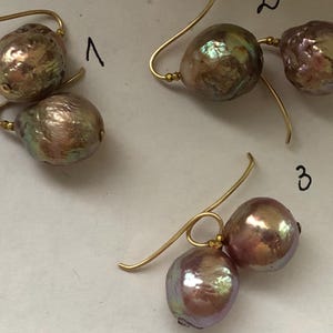 May include: Three pairs of earrings with iridescent, baroque-shaped pearls. Each earring features a gold-coloured wire hook. The pearls display a mix of colours, including pink, gold, and green, with a lustrous sheen. The earrings are displayed on a white surface.