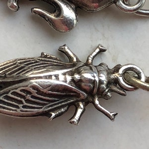 May include: A silver charm in the shape of a cicada insect. The charm has detailed wings and legs, and a loop for attaching to a bracelet or necklace.