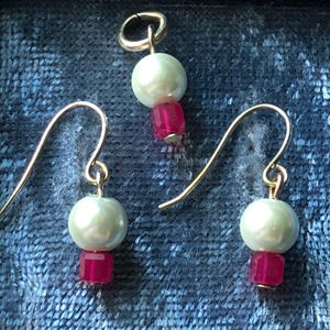 May include: A jewelry set featuring a pendant and two earrings. Each piece has a pale green pearl-like bead above a small, square, magenta-colored bead. The earrings and pendant have gold-colored hooks and loops.