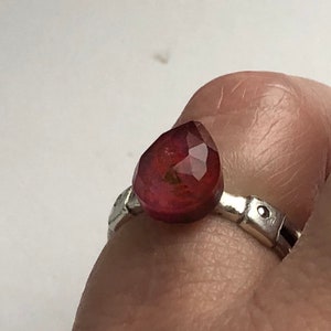 May include: A silver ring with a teardrop-shaped, faceted, deep red gemstone.