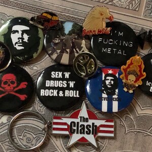 May include: A collection of assorted pins and badges with rock and roll, political, and pop culture themes. Includes a Che Guevara pin, a skull and crossbones badge, and a pin with the text "I'm Fucking Metal".