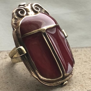 May include: A gold-toned ring featuring a large, reddish-brown scarab beetle design. The ring has an ornate, detailed setting with a rectangular shape and a polished, glossy finish. The ring's band is also gold-toned.