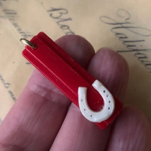 May include: A red plastic charm with a white horseshoe design. The charm has a gold loop for attaching to a necklace or bracelet.