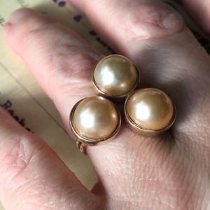 May include: A gold-toned ring featuring three large, round, cream-colored faux pearls. The pearls are set in individual gold bezels, creating a cluster design. The ring is shown on a hand.