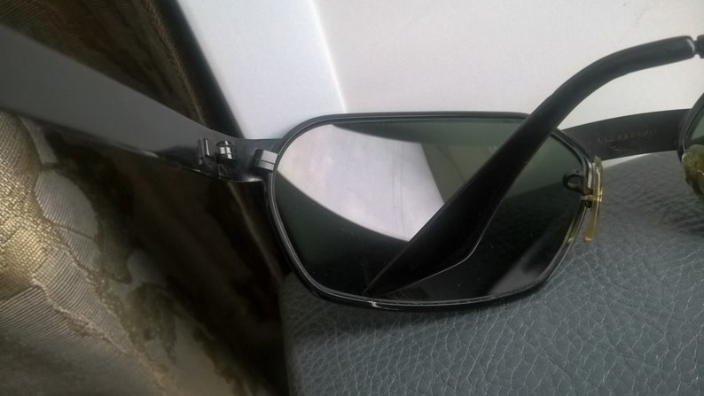 May include: Black sunglasses with a dark green lens and a gold accent on the side. The sunglasses are made of plastic and have a classic aviator style.