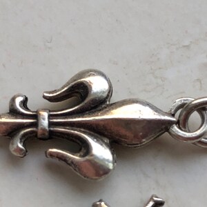 May include: Silver charm featuring a fleur-de-lis design with a loop for attaching to a bracelet or necklace.