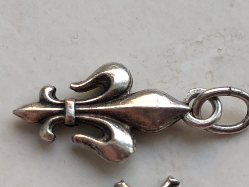 May include: Silver charm featuring a fleur-de-lis design with a loop for attaching to a bracelet or necklace.