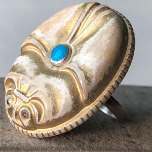 May include: A silver-toned ring with an oval, gold-colored face. The face features an embossed design with a turquoise stone and a small, clear stone. The ring has a textured edge and a simple band.