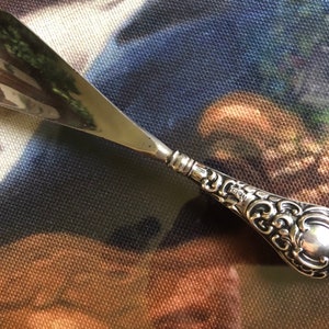 May include: A silver shoehorn with an ornate, detailed handle. The handle is decorated with a floral design and has a smooth, rounded end.