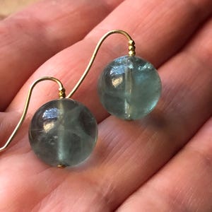 Op de afbeelding: Een paar goudkleurige oorhangers met ronde, doorschijnende, lichtblauwgroene kralen. De oorbellen hebben een eenvoudig, elegant ontwerp met een haaksluiting. De kralen hebben een diameter van ongeveer 1,3 cm.