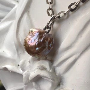 以下が含まれることがあります： シルバーチェーンのネックレス。大きな、不規則な形をした、虹色の茶色の真珠のペンダントが付いています。真珠は質感のある表面で、複数の色を反射します。ネックレスは白い彫刻された表面に展示されています。