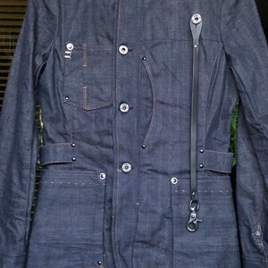 May include: A dark blue denim jacket with a long black strap and multiple pockets. The jacket has a stand-up collar and a button closure.