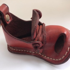 Puede incluir: Un pequeño llavero de cuero marrón en forma de zapato con un anillo para llaves adjunto. El zapato tiene una solapa que se abre para revelar un pequeño bolsillo en el interior.