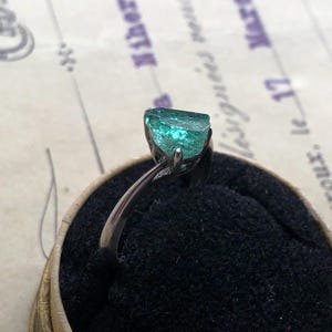 Könnte beinhalten: Ein Silberring mit einem rohen, teal-farbenen Edelstein. Der Ring ist auf einem schwarzen Samthintergrund in einer kleinen, runden Schachtel präsentiert. Der Hintergrund zeigt verblassten Text auf gealtertem Papier.