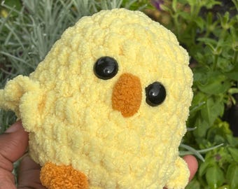 May include: A small, round, yellow crocheted chick plush toy with a textured, fuzzy yarn surface. It features two large, round, black plastic safety eyes, a small orange beak, and two small orange feet made from the same textured yarn material.