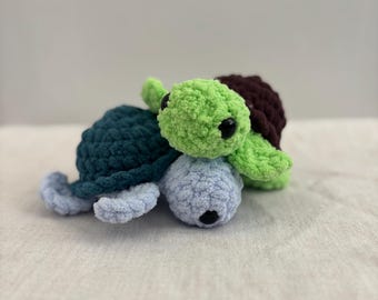 May include: Three crocheted turtles in shades of green, blue, and brown. The turtles have black eyes and are made of a soft, textured yarn. The turtles are arranged together, creating a playful and whimsical scene.
