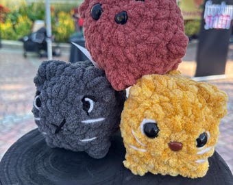 May include: Three handmade crochet cat toys in shades of red, gray, and yellow. Each toy has black button eyes and white whiskers. The toys are displayed on a black wooden surface.