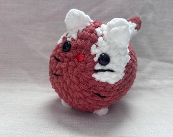 May include: A crocheted plush toy cat with a red and white color scheme. The cat has black eyes and a black nose.