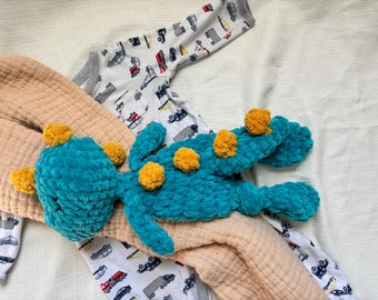 May include: A teal blue crocheted dinosaur toy with yellow pom-poms. The toy is lying on a beige blanket with a white and grey patterned baby onesie.