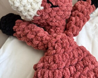 May include: A crocheted plush fox toy with a red body, white face, and black eyes. The toy is lying on a white surface.