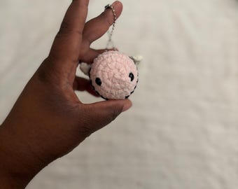 May include: A handmade crochet bee keychain. The bee is primarily light pink with black and white accents. It has small white wings and a silver chain for attachment. The bee is a small, round shape.