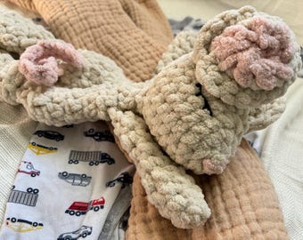 May include: A beige crocheted plush toy shaped like a bunny with pink ears. The bunny is lying on a beige blanket.