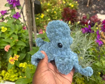 May include: A small, blue, crocheted octopus plush toy with a textured, soft yarn surface and two dark, round, glossy plastic eyes. The octopus features a rounded head and several curled, tentacle-like arms extending from the base.