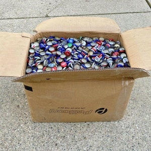 May include: A cardboard box overflowing with a large collection of colourful bottle caps. The box is labelled "Pepsi-Cola" and "On Top Of The Best".
