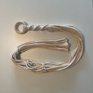 May include: A white macrame plant hanger with a wooden ring at the top. The hanger is made of multiple strands of rope that are knotted together.