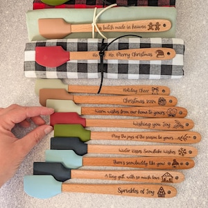 May include: A collection of wooden-handled silicone spatulas with engraved holiday messages and festive icons. Three spatulas are tied to folded plaid and solid-colored kitchen towels, while others are arranged below with various seasonal phrases.