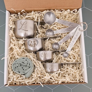 May include: A set of stainless steel measuring cups and spoons in a cardboard box with paper filler. The cups feature engraved letters and a couple silhouette. The spoons are on a ring with the text Love is the secret ingredient, Josh & Breanna, 10-18-2025.