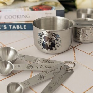 May include: A set of stainless steel measuring cups and spoons featuring engraved text and illustrations. The cups display a silhouette of a couple within a floral wreath, while the spoons are marked with measurements and the names Logan & Serina.