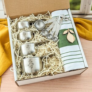 May include: A gift box containing four stainless steel measuring cups with floral engravings, a set of four stainless steel measuring spoons, and a white kitchen towel with green stripes and a wooden lemon-shaped ornament attached to the fabric.