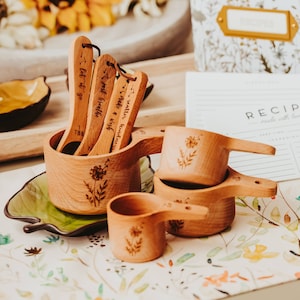 May include: Wooden measuring cups and spoons with floral designs. The spoons have inspirational phrases like "Let it go" and "Scatter kindness." A recipe card and a floral-patterned container are in the background. The measuring spoons are marked with "1 TBS".