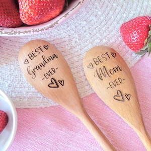 May include: Two light brown wooden spoons with laser-engraved text and heart symbols. One spoon features the text BEST Grandma -EVER- and the other features the text BEST Mom -EVER-. The spoons are positioned near fresh red strawberries.