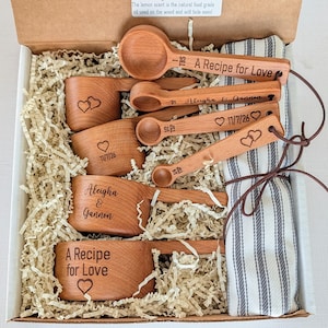May include: A set of wooden measuring cups and spoons in a cardboard box with a striped cloth. The items are engraved with the text A Recipe for Love, Aleigha & Gannon, 11/7/26, and heart symbols. The spoons include 1 TBS, 1 TSP, 1/2 TSP, and 1/4 TSP.